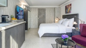 A modern hotel room with a bed, bedside table, a small round table with a vase, a red chair, a TV on the wall, and a kitchen area with a counter and appliances.