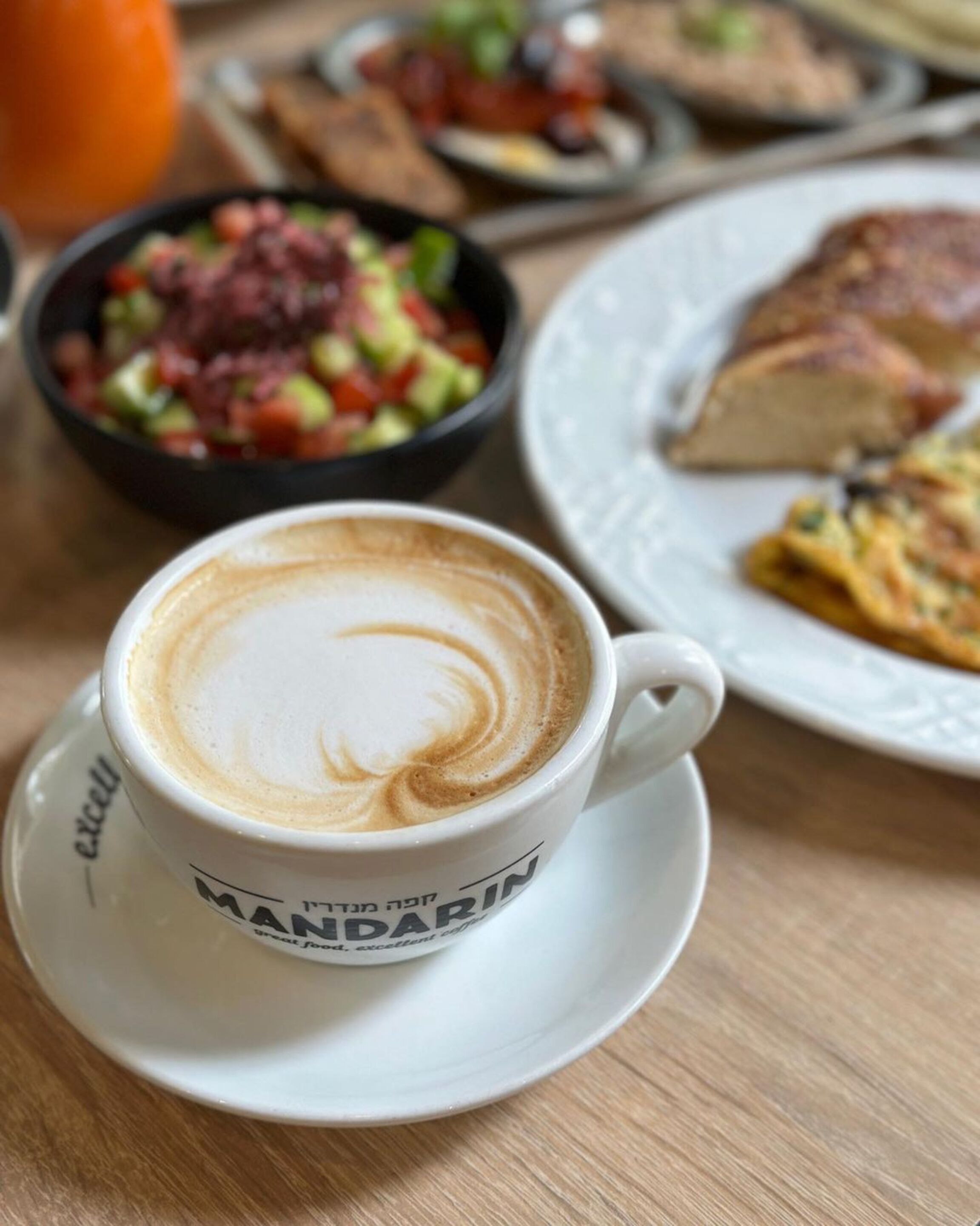A cup of coffee with latte art on a saucer, a plate with a slice of cake, and a bowl of salad.