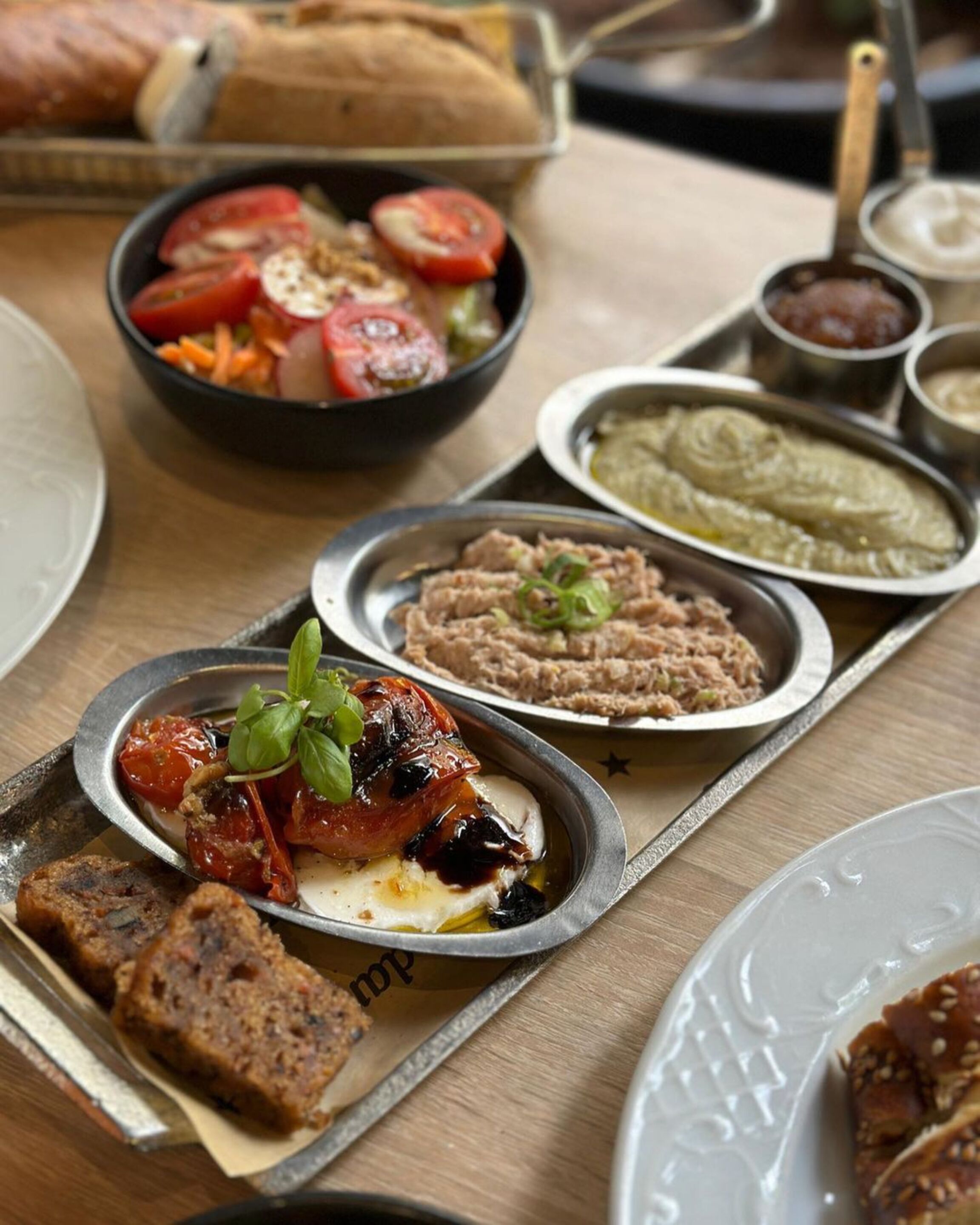 Variedad de platos, incluyendo un plato con tomates a la parrilla y mozzarella, una ensalada en un bol, y varios recipientes pequeños con salsas.