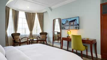 Habitación de hotel con una cama grande, dos sillones, un escritorio con lámpara, un espejo y un cuadro de un edificio en la pared.