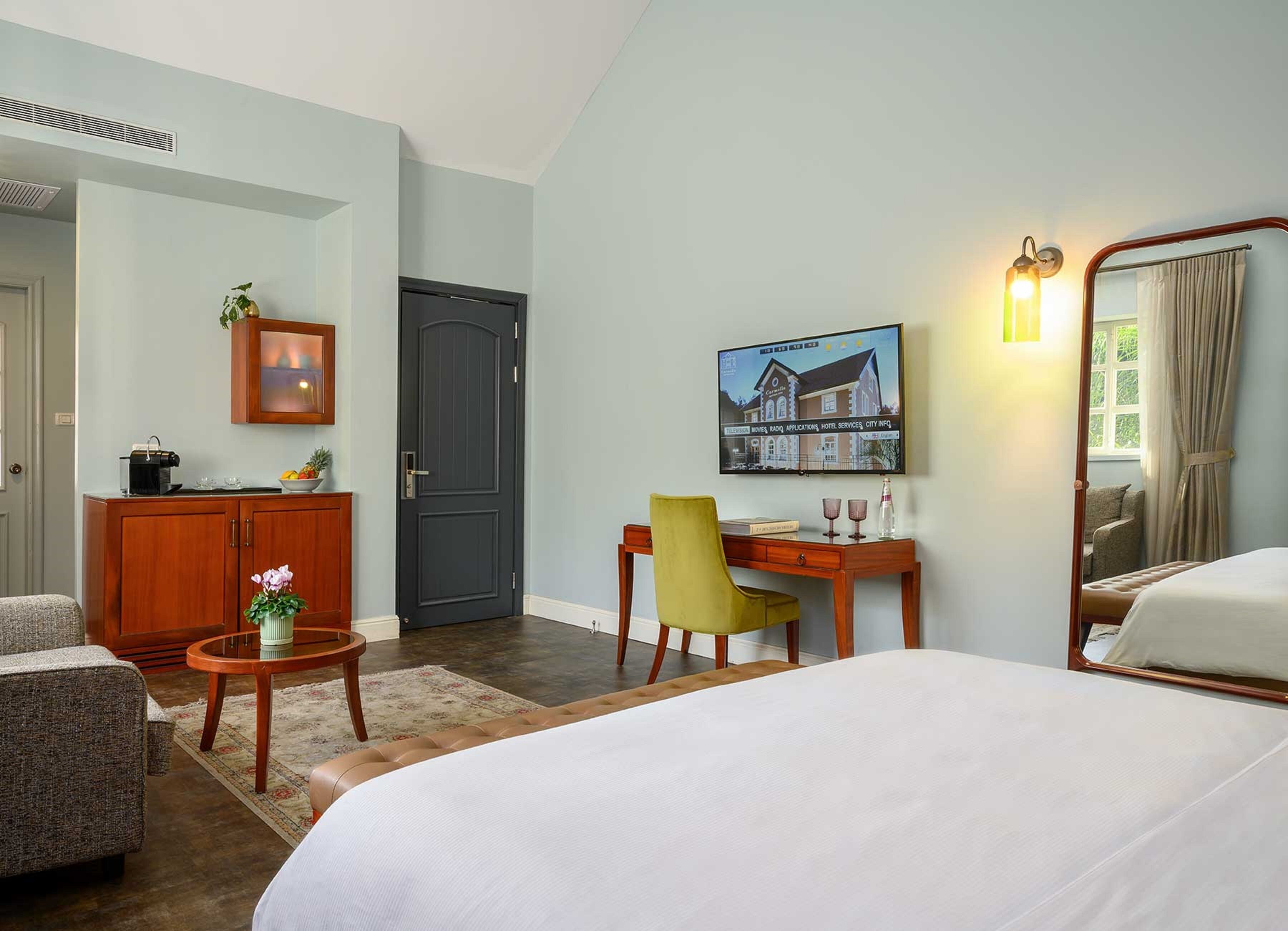 A hotel room with a bed, a desk, a chair, a small table with a plant, a cabinet, a television, and a framed picture on the wall.