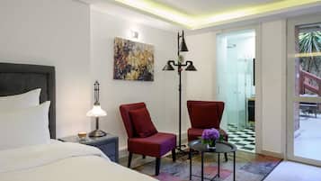 A hotel room with a bed, two red chairs, a floor lamp, a table with a vase, and a painting on the wall.