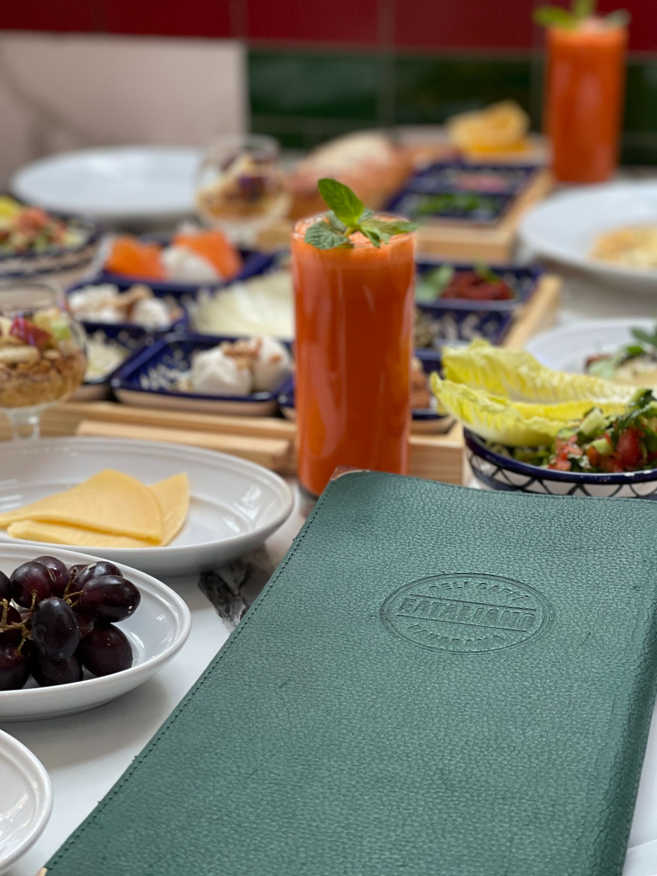 A table set with various dishes, including a tall glass of tomato juice, cheese, grapes, and a green menu.