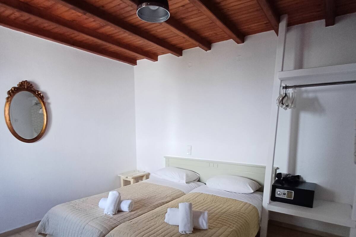 A double bed with beige bedding and white towels, a wooden ceiling, a white wall, a round mirror, and a bedside table with a radio.
