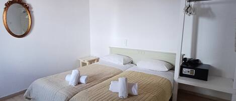 A double bed with beige bedding and white towels, a wooden ceiling, a white wall, a round mirror, and a bedside table with a radio.