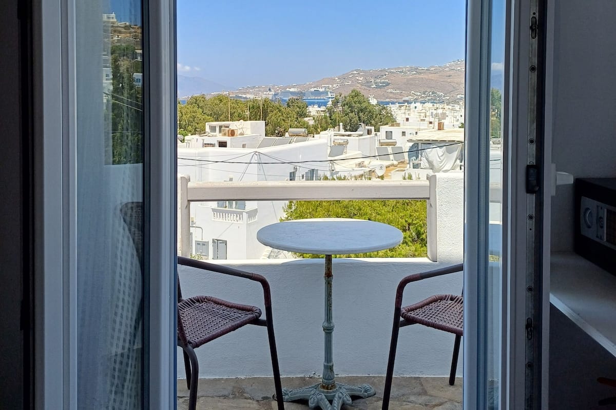 A balcony with two wicker chairs and a small round table, overlooking a cityscape with buildings and a clear blue sky.