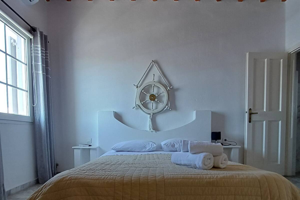 A bedroom with a wooden ceiling, a bed with a beige quilt, a wall-mounted fan, and a door to the right.