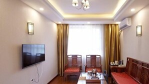 Una habitación de hotel con televisión de pantalla plana en la pared, una mesa de madera con sillas, un sofá y un ventanal con cortinas.
