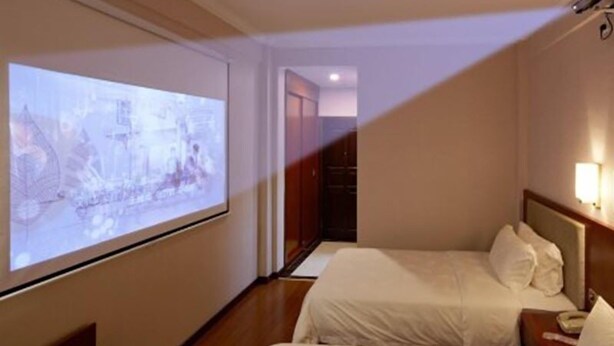 Una habitación de hotel con una cama, una mesita de noche, una pantalla proyectora mostrando un paisaje urbano y un armario.