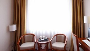 Habitación de hotel con dos sillas de madera, una mesita con una bandeja de objetos, un escritorio con una computadora y una ventana con cortinas.