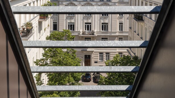 A view from a window overlooking a row of classical buildings with balconies, a clear sky, and a few cars on the street below. - Rafael Kaiser - Premium Apartments City Centre  (Vienna)