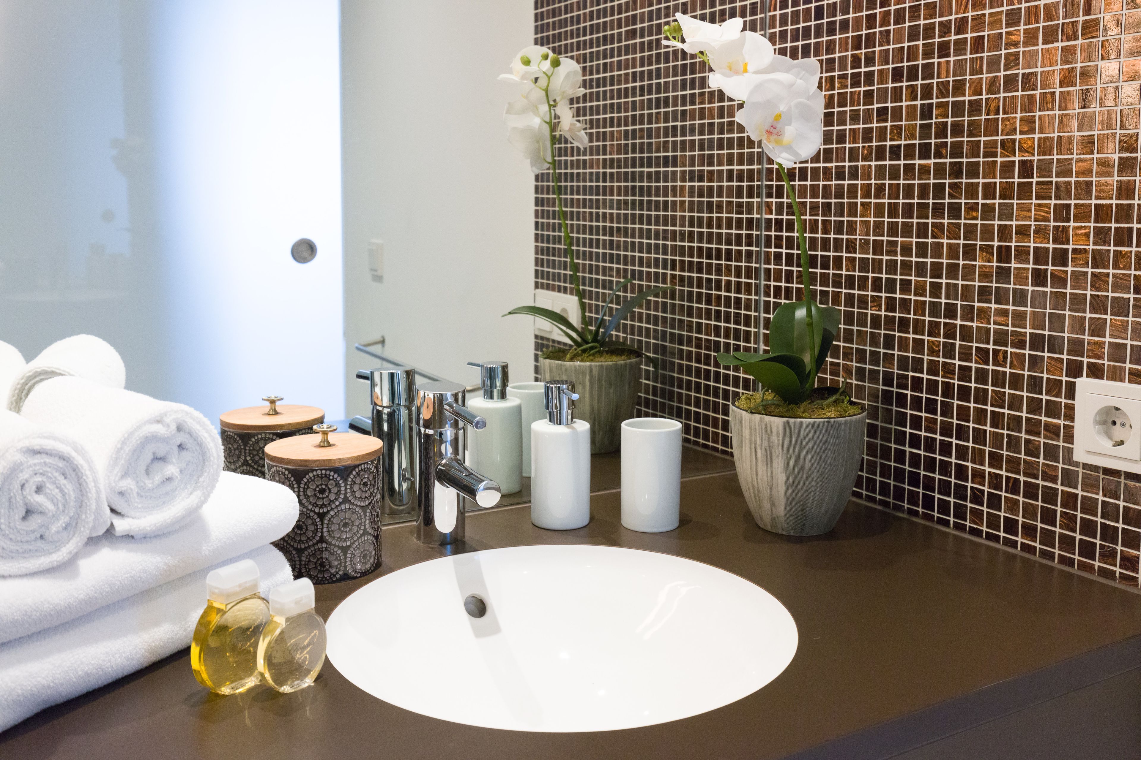 A modern bathroom with a round white sink, a mirror, and a tiled wall. There are towels stacked on the left, a potted orchid on the right, and a small plant in a pot on the counter.