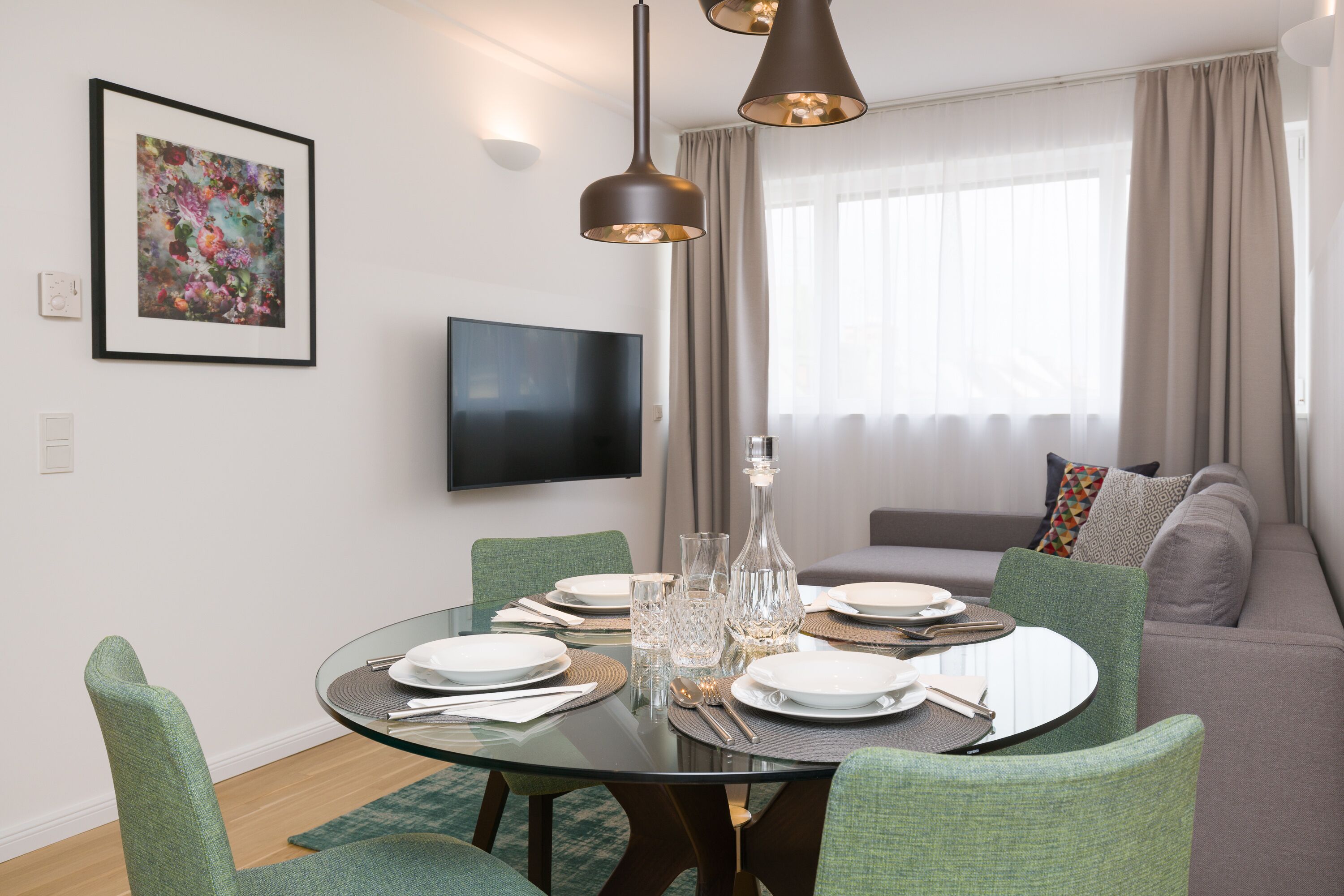 Un comedor moderno con una mesa de vidrio, sillas tapizadas de verde, un televisor de pantalla plana y un ventanal con cortinas.