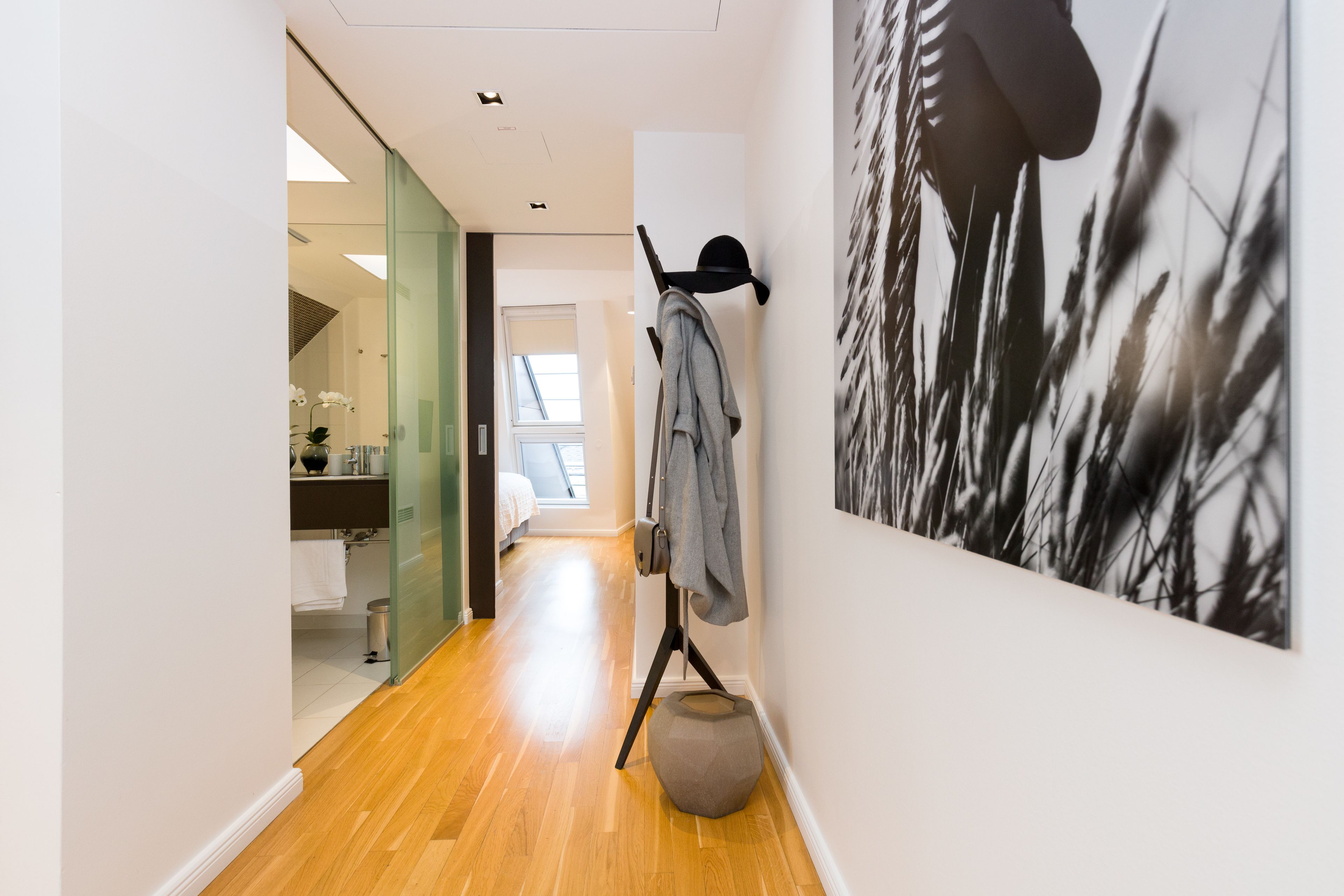A hallway with a wooden floor, a coat rack with a jacket, a round seating area, and a large black and white artwork on the wall.