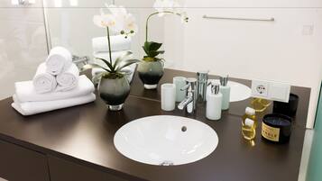 Ein modernes Badezimmer mit einem großen weißen Waschbecken, einem Spiegel und einem Handtuchhalter. Auf der Ablagefläche befinden sich zwei Orchideen in Töpfen und eine Kerze.