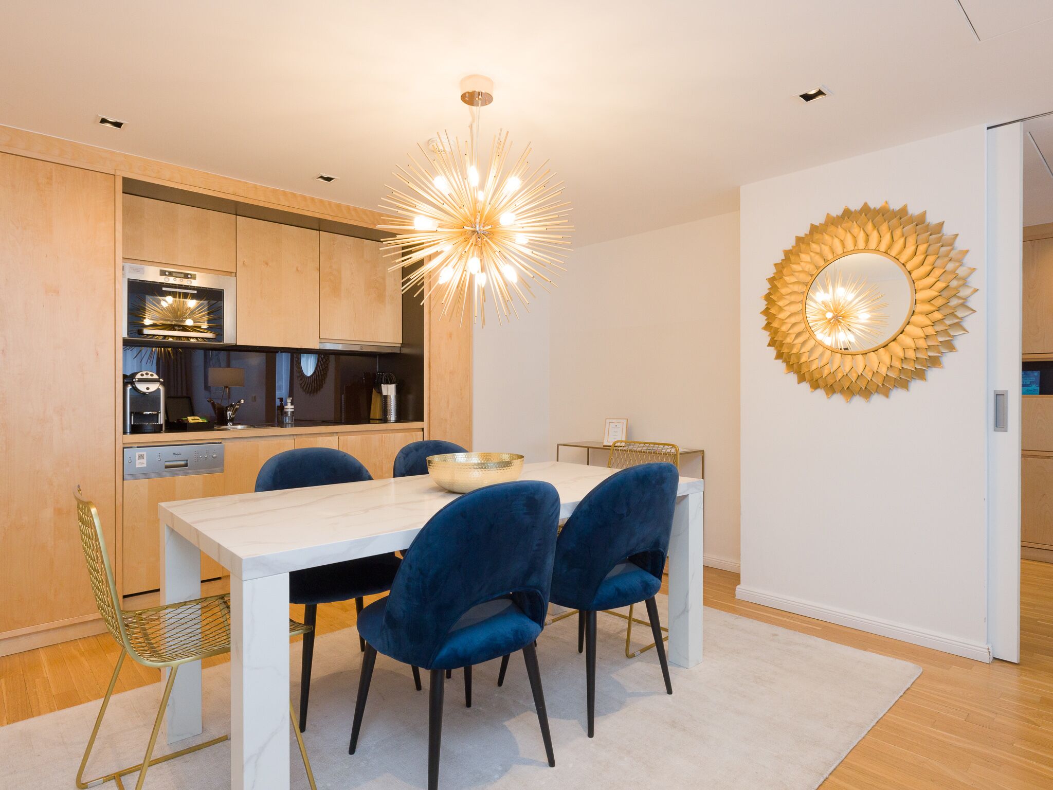A modern kitchen with wooden cabinets, a built-in oven, and a dining area with a white table and blue chairs.