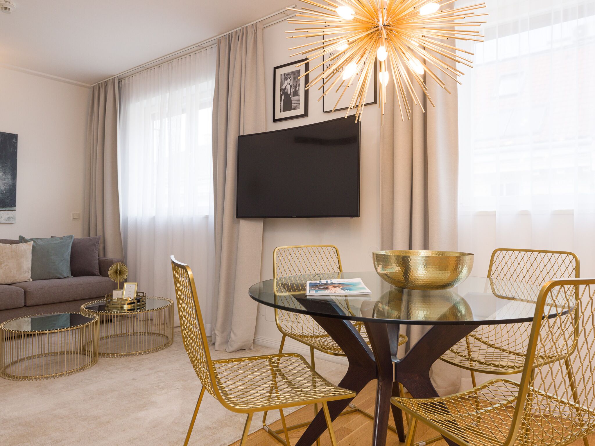 Een moderne woonkamer met een glazen eettafel, gouden draadstoelen, een salontafel met gouden lijst en een grote flatscreen-tv, plus een decoratieve lamp.