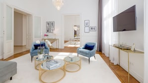 A modern living room with a chandelier, two armchairs, a coffee table, and a flat-screen TV mounted on the wall. - Rafael Kaiser - Premium Apartments City Centre  (Vienna)
