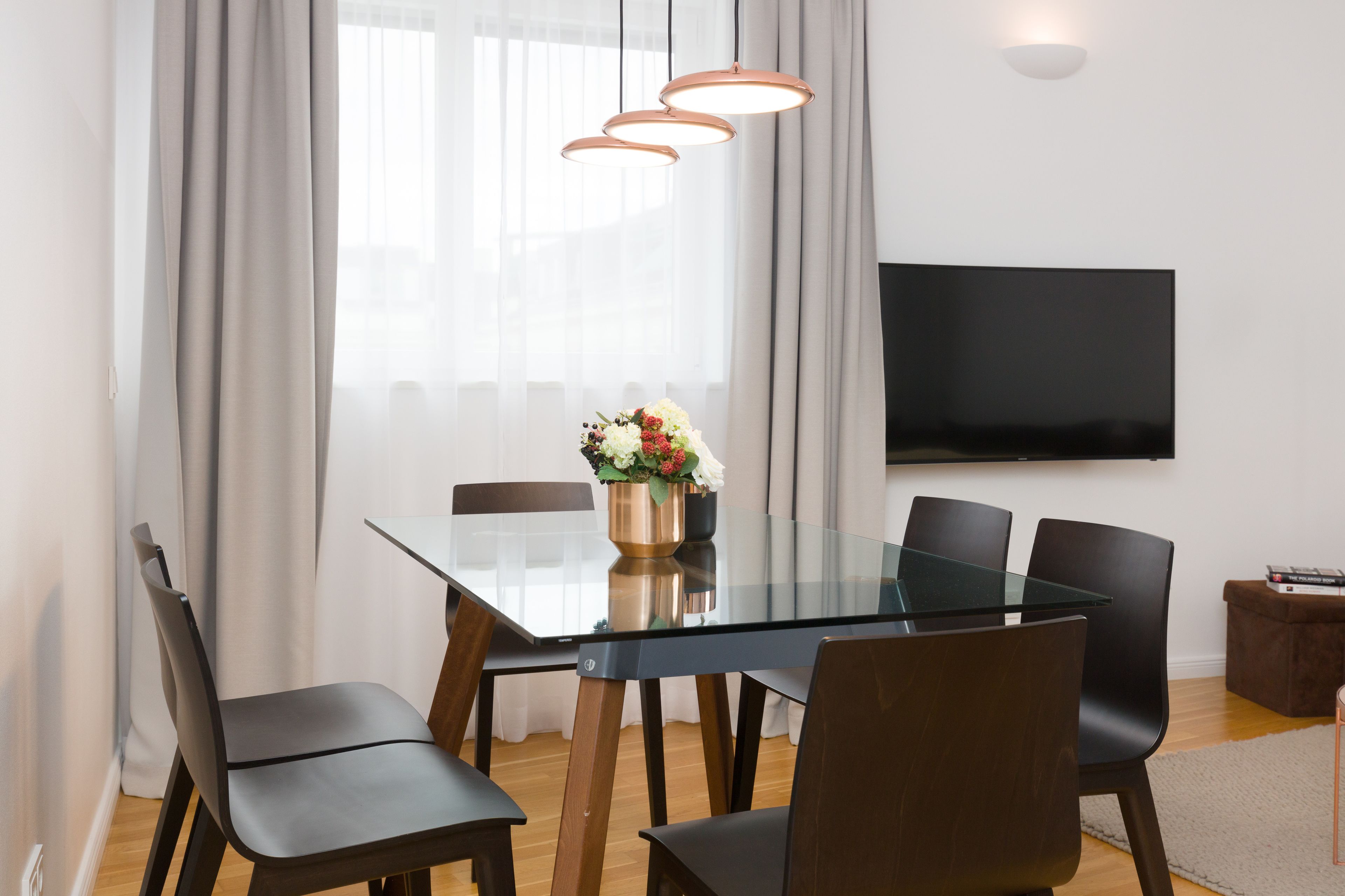 A modern dining area with a glass table, black chairs, a vase of flowers, and a flat-screen TV on the wall.