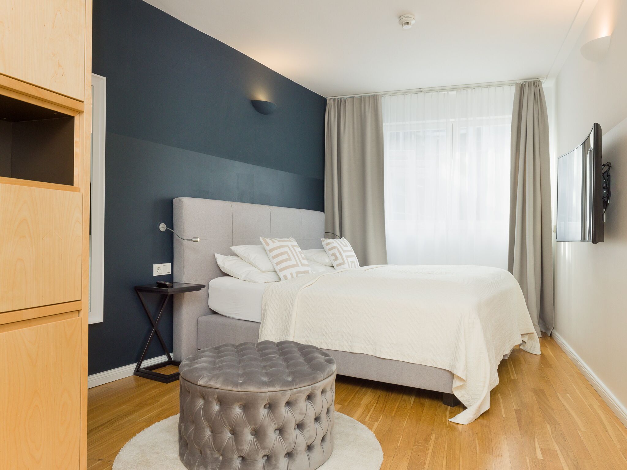 A modern bedroom with a large bed, a grey ottoman, a nightstand, and a wardrobe.