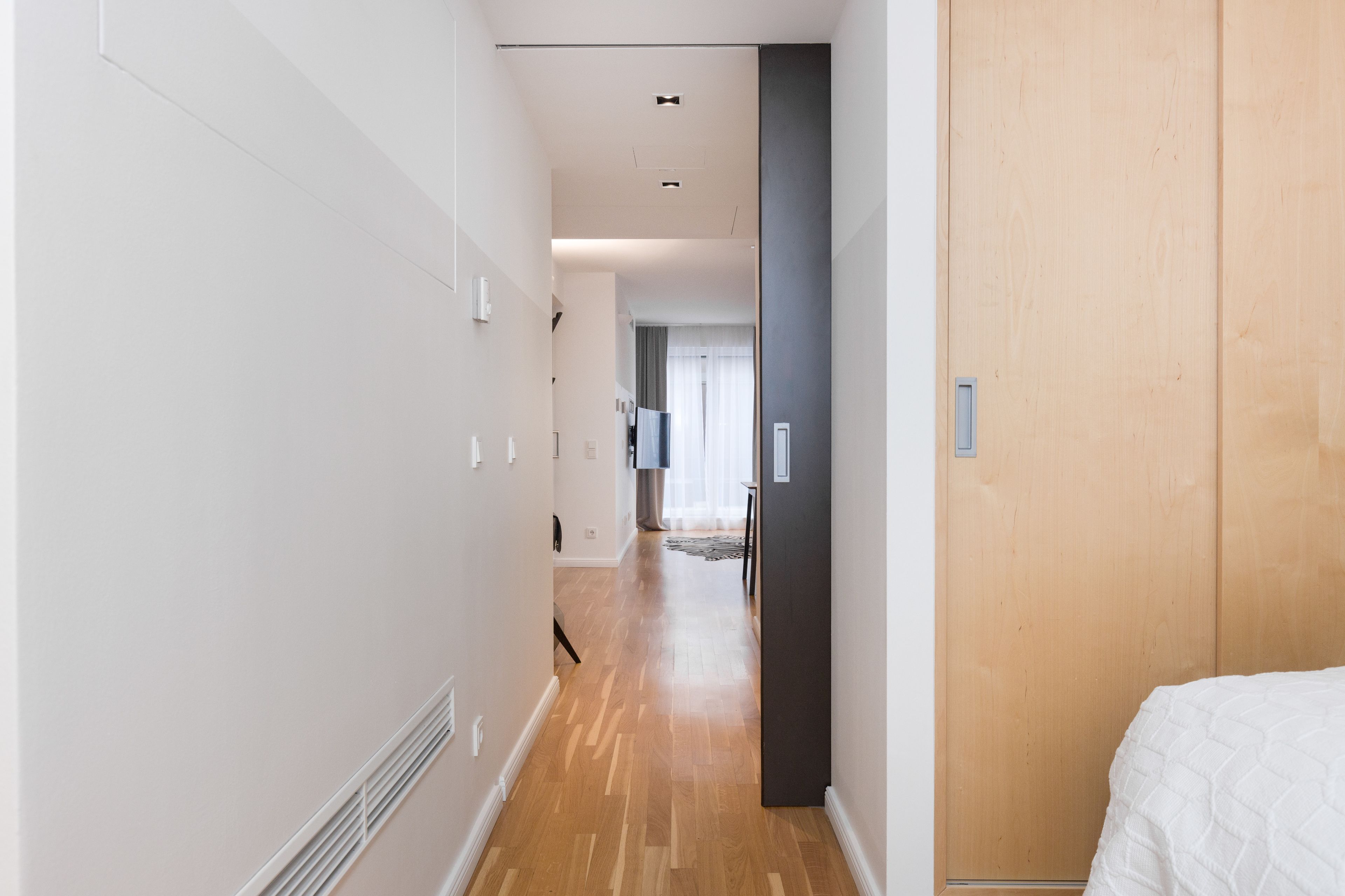 A narrow hallway with wooden flooring and white walls, leading to a room with a bed.