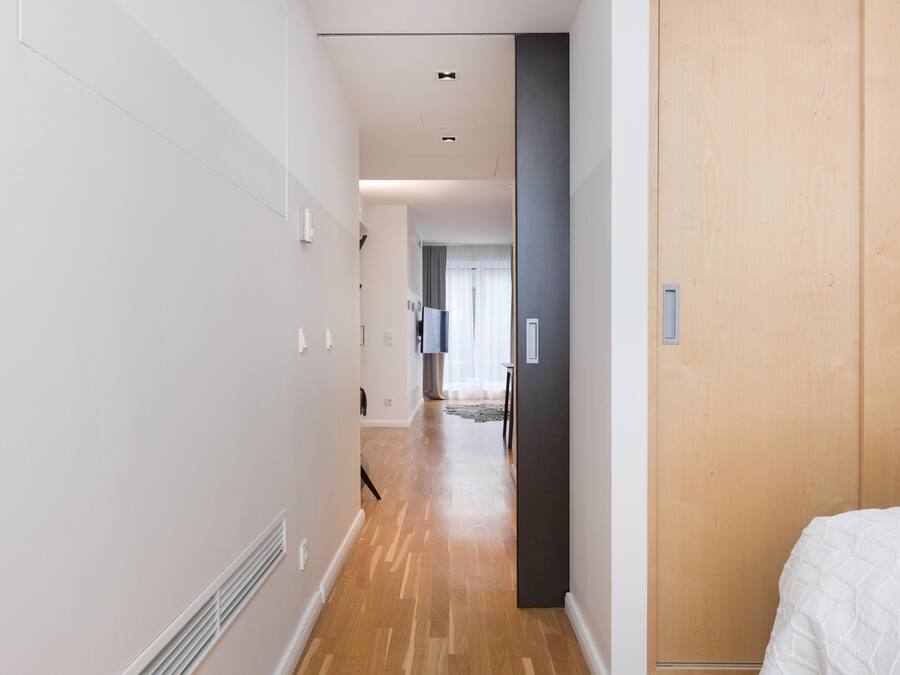 A narrow hallway with wooden flooring and white walls, leading to a room with a bed.