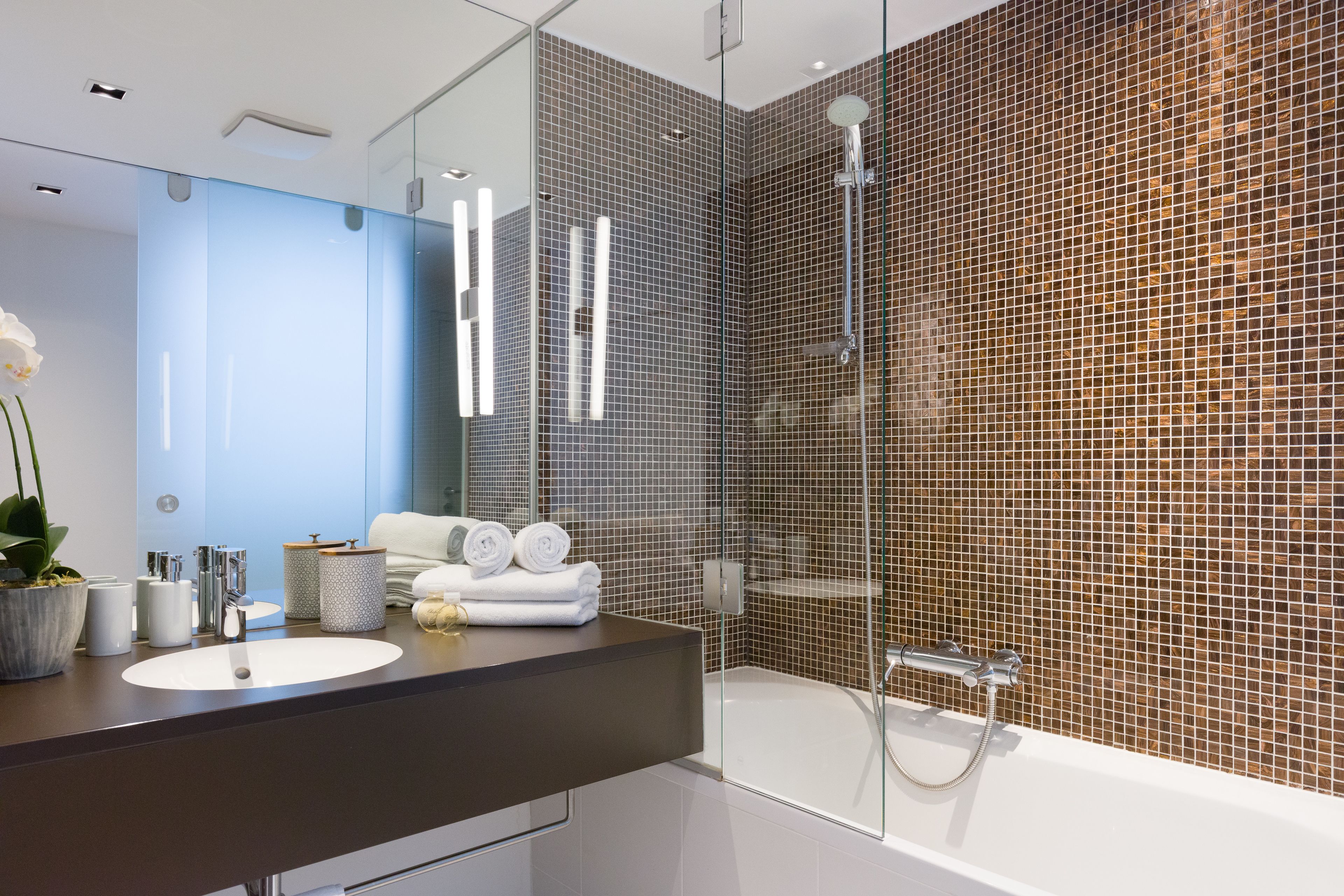 Un baño moderno con cabina de ducha de vidrio, un espejo grande y una encimera oscura con lavamanos y artículos de aseo.