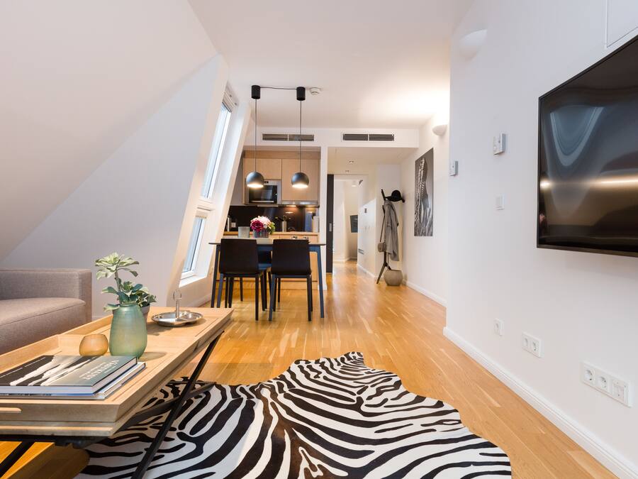 A modern living room with a sofa, a wooden coffee table with books and a vase, a zebra-patterned rug, a kitchen in the background, and a TV mounted on the wall.