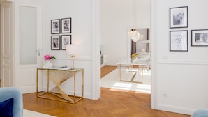 A modern living room with a glass coffee table, a gold console table, and a blue sofa. There are framed pictures on the walls and a chandelier hanging above the dining table. - Rafael Kaiser - Premium Apartments City Centre  (Vienna)