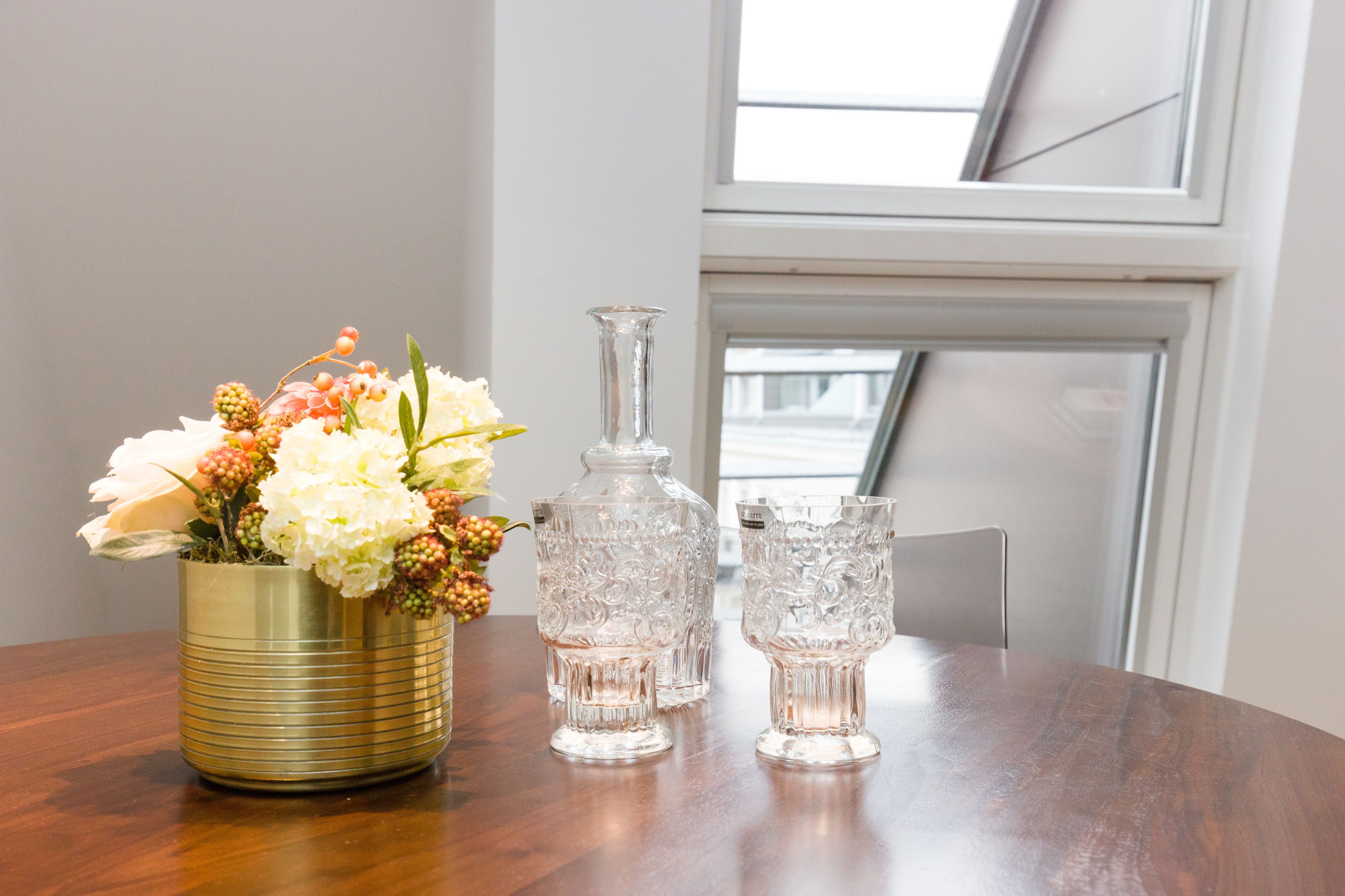 Una mesa con un jarrón dorado con flores, una jarra de cristal transparente y dos copas de cristal.