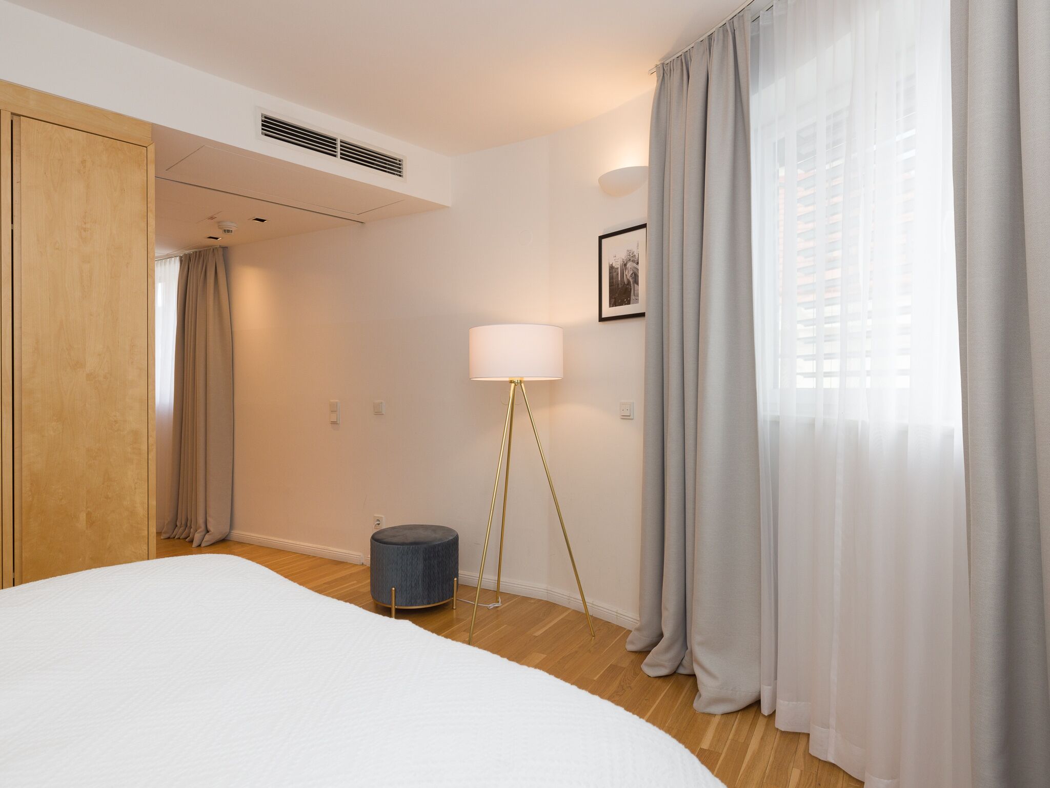 A hotel room with a bed, a standing lamp, a round blue ottoman, and a window with sheer curtains.