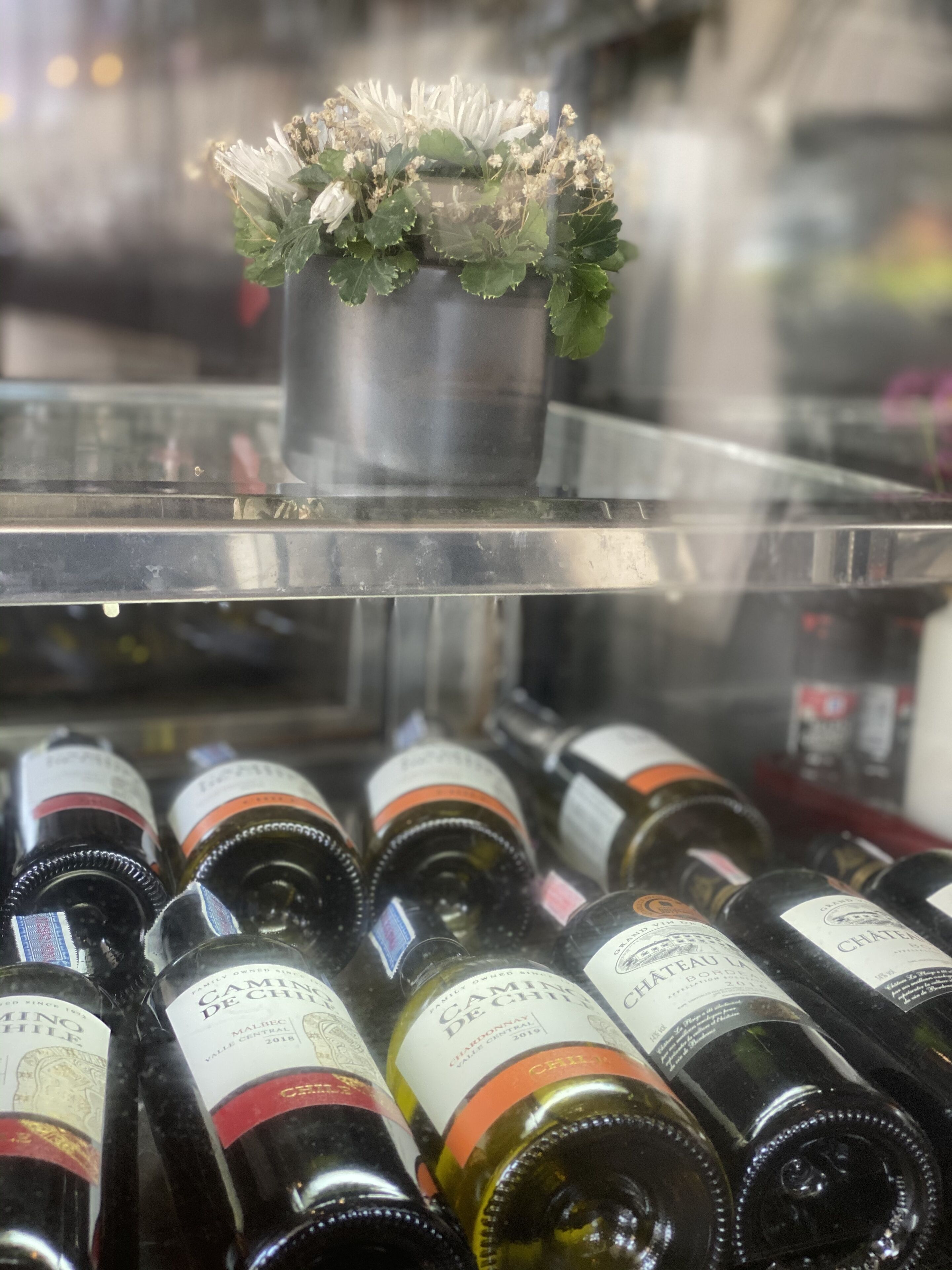 Una exhibición de botellas de vino con etiquetas, un recipiente metálico con flores y una barrera de cristal.