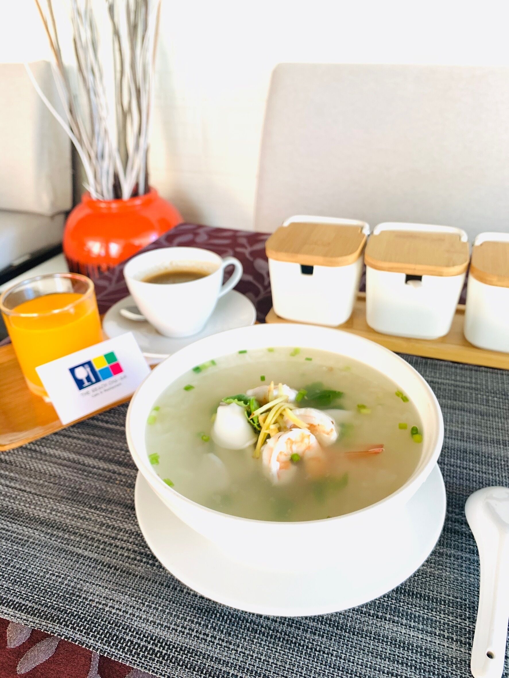 A bowl of soup with shrimp and vegetables, a cup of coffee, a glass of orange juice, and a small orange vase with white branches.