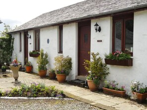 A white building with a dark brown door and windows, potted plants, and a stone pathway. - Shegarton Farm Cottages (Alexandria)