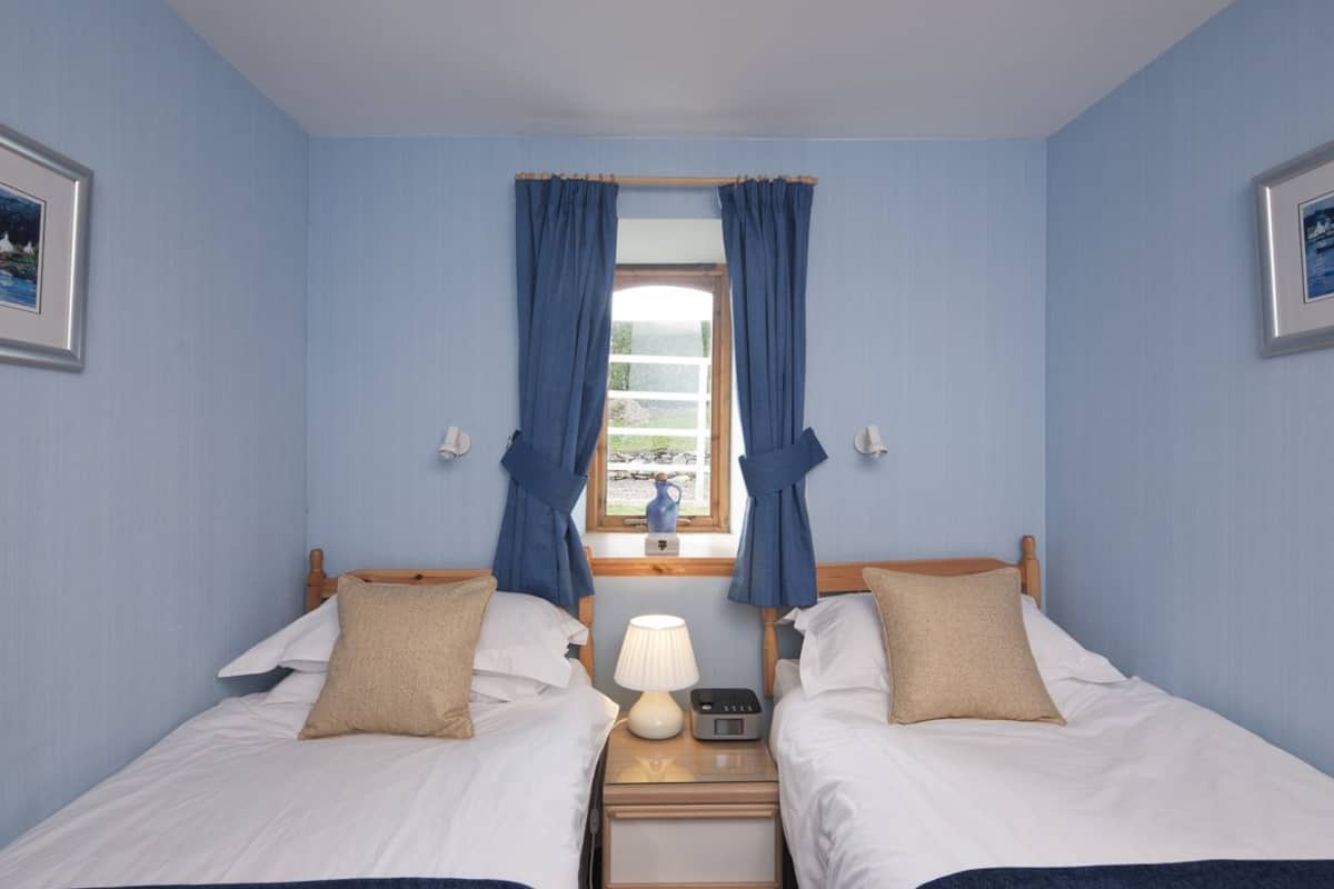 A room with two beds, a nightstand with a lamp, a window with curtains, and wall-mounted light fixtures.