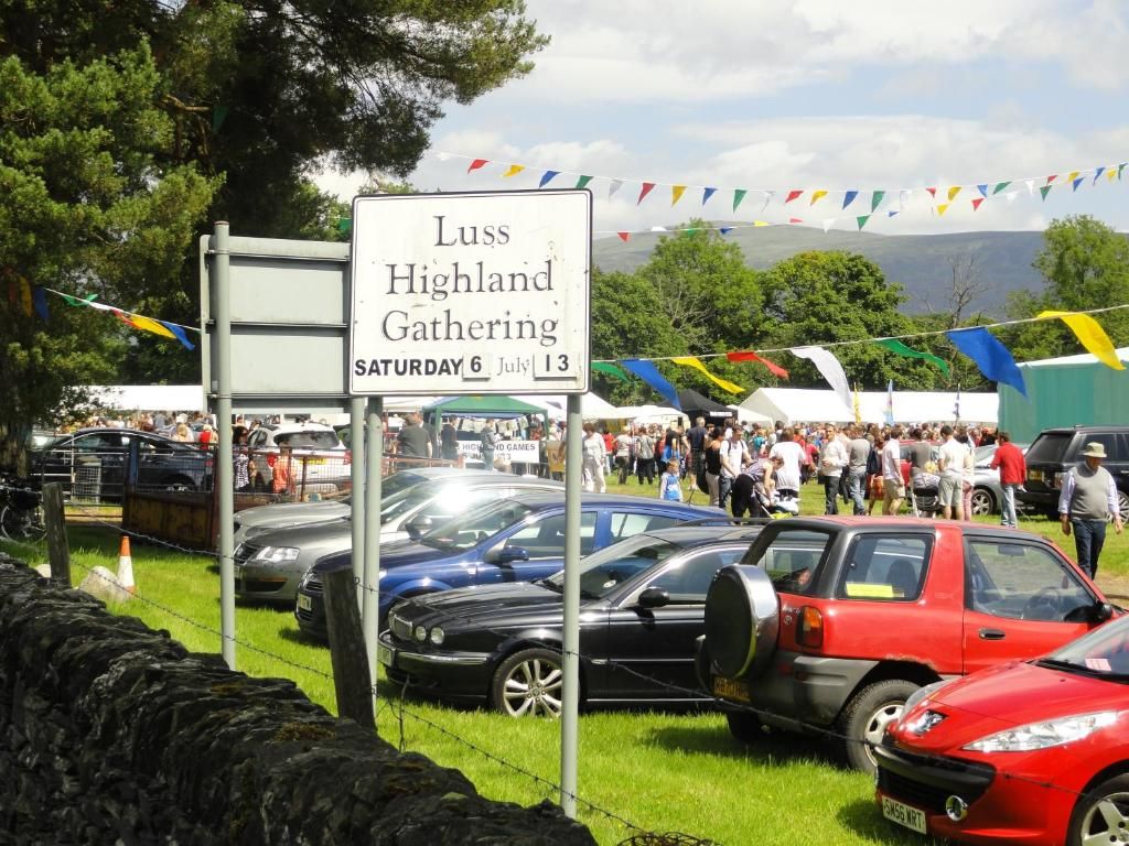 Une pancarte annonçant le festival de Luss Highland Gathering, visible un samedi, entourée de voitures garées et d’une foule de personnes.