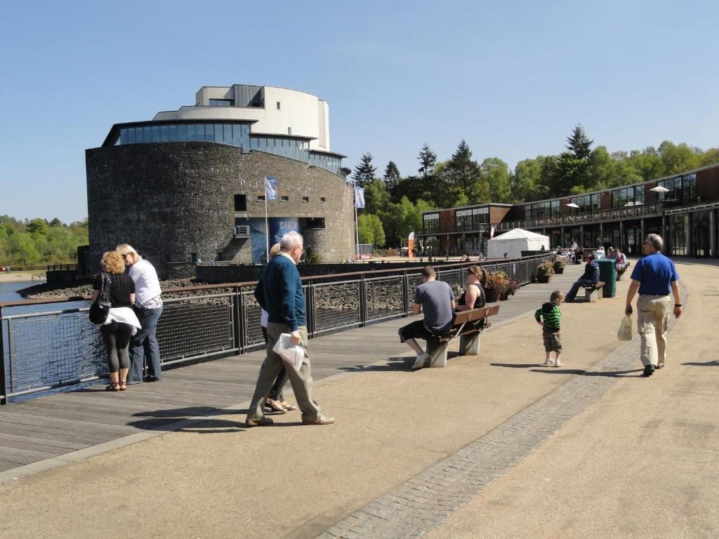 A waterfront promenade with people walking, sitting on benches, and a modern building with a circular section.