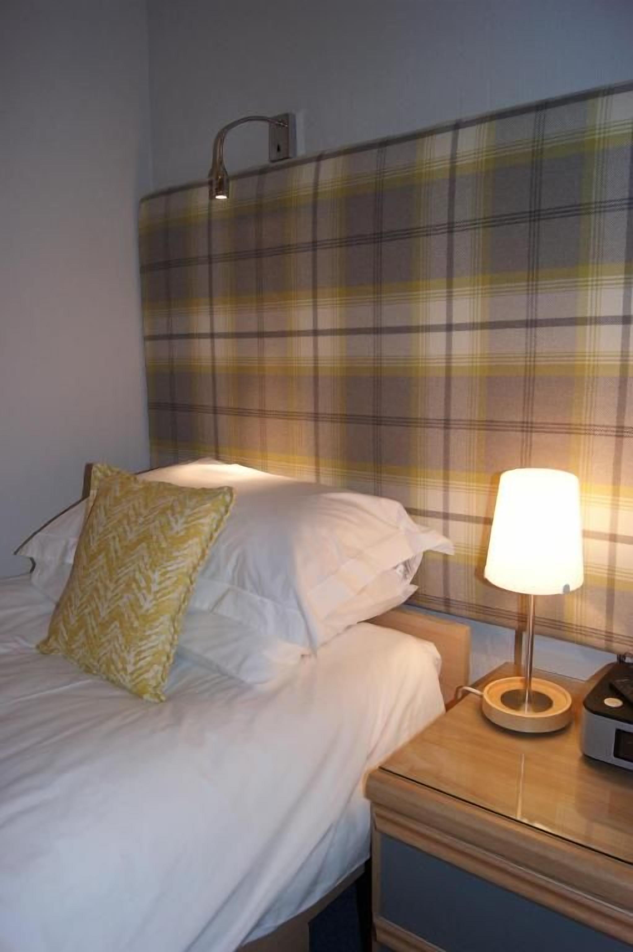 A bed with white bedding and a yellow pillow, a bedside table with a lamp, and a plaid-patterned headboard.