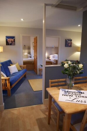 A hotel room with a bed, a blue sofa, a dining table with chairs, and a vase with flowers. - Shegarton Farm Cottages (Alexandria)