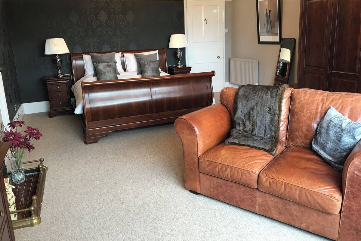 Une chambre à coucher avec un grand lit, un canapé en cuir marron, une table de chevet, une commode et un lustre.