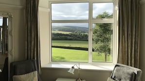 A room with a large window offering a view of a green landscape and hills. There is a small table with a lamp and two chairs. - Cedar House (Minehead)