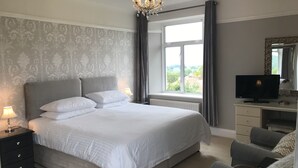 A bedroom with a large bed, a desk with a TV, a chair, and a window with a view. - Cedar House (Minehead)
