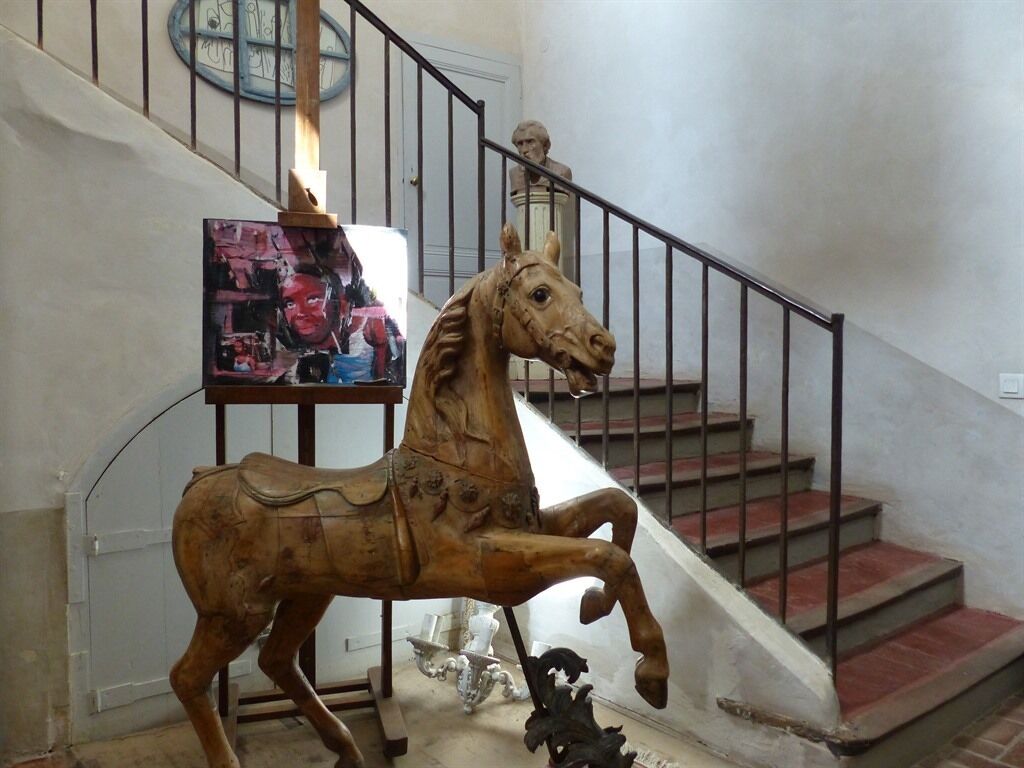 A wooden horse sculpture in a hallway with a staircase and a painting on the wall.