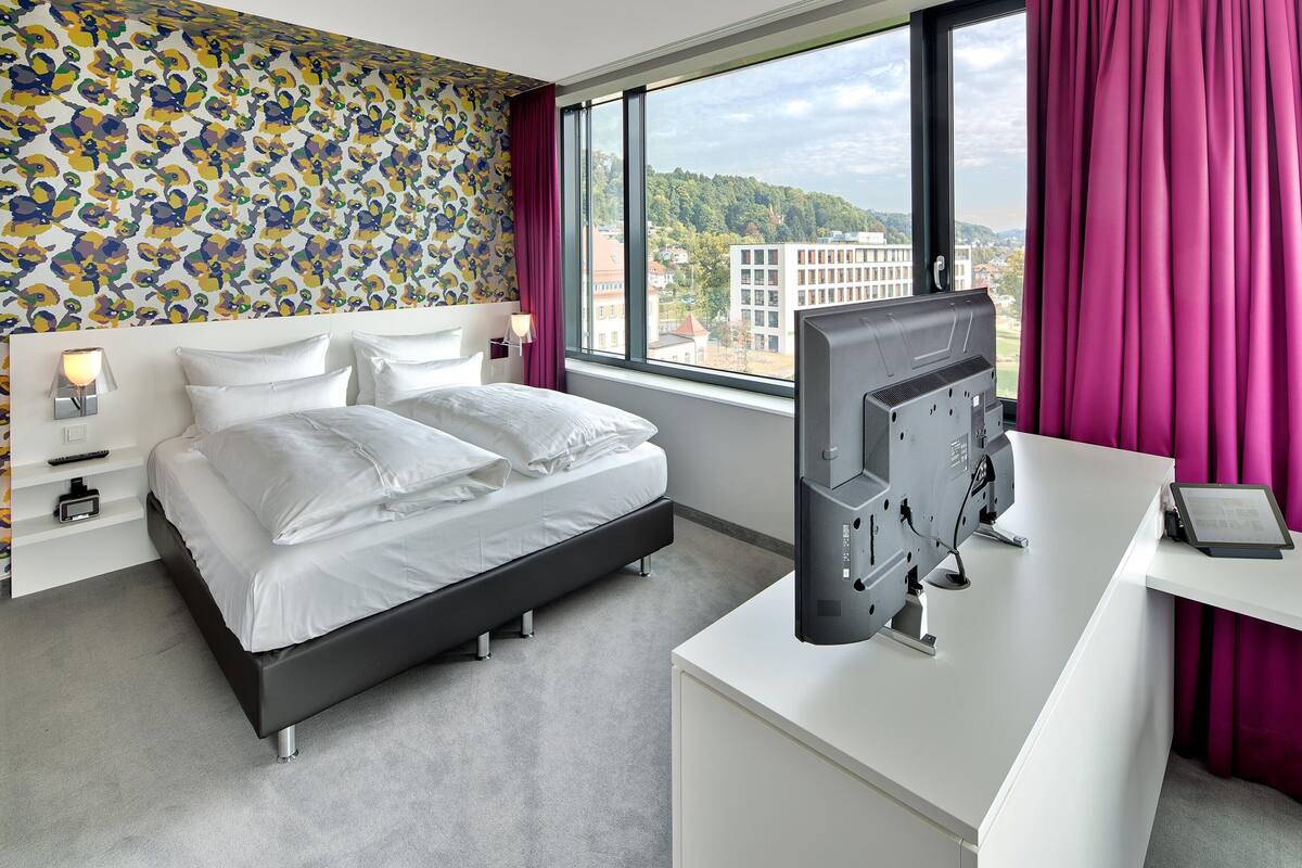 Ett modernt hotellrum med en säng, ett skrivbord med en dator, en TV och ett stort fönster med utsikt över byggnader och kullar.