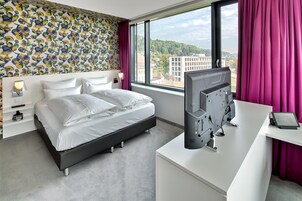 Una habitación de hotel moderna con una cama, un escritorio con una computadora, un televisor y un amplio ventanal con vistas a edificios y colinas.