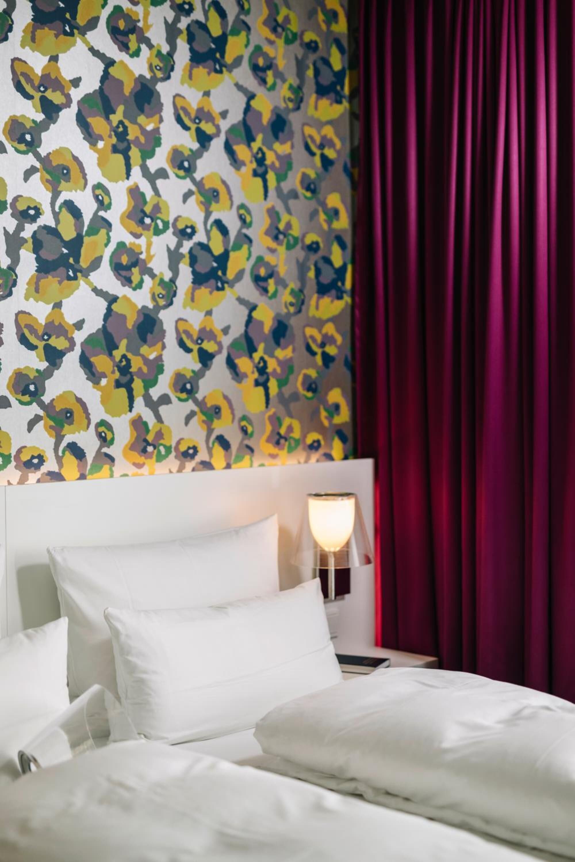 A hotel room with a bed, white pillows, a nightstand with a lamp, and a wall with a floral pattern.