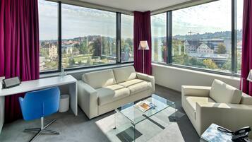A modern hotel room with a large window, a sofa, a glass coffee table, and a desk with a chair.