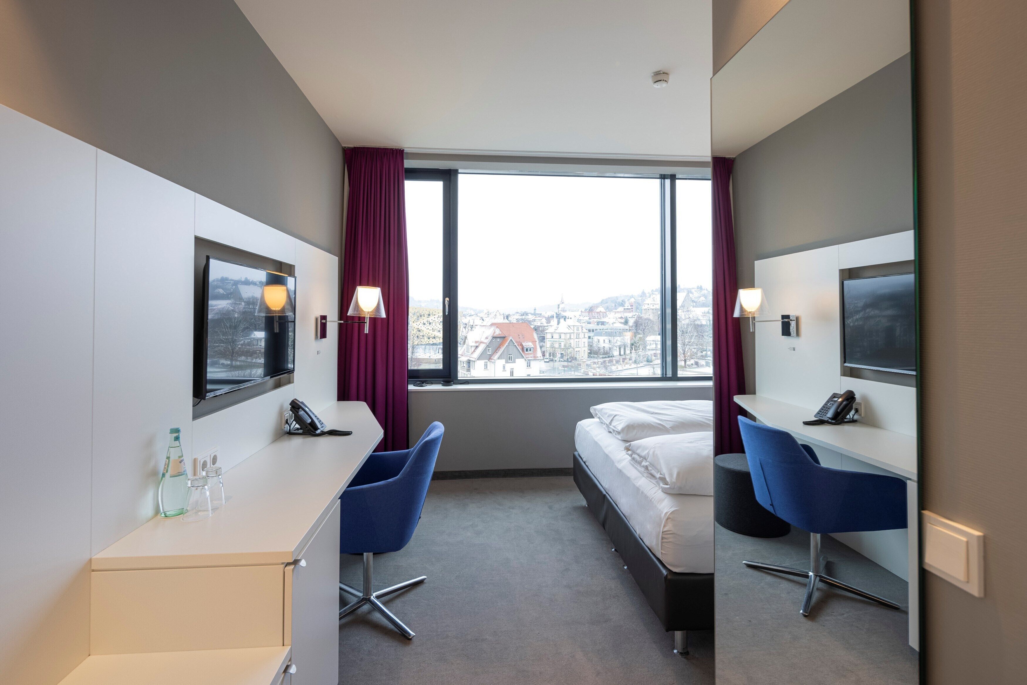 A hotel room with a bed, two blue chairs, a desk, a TV, and a window with a city view.