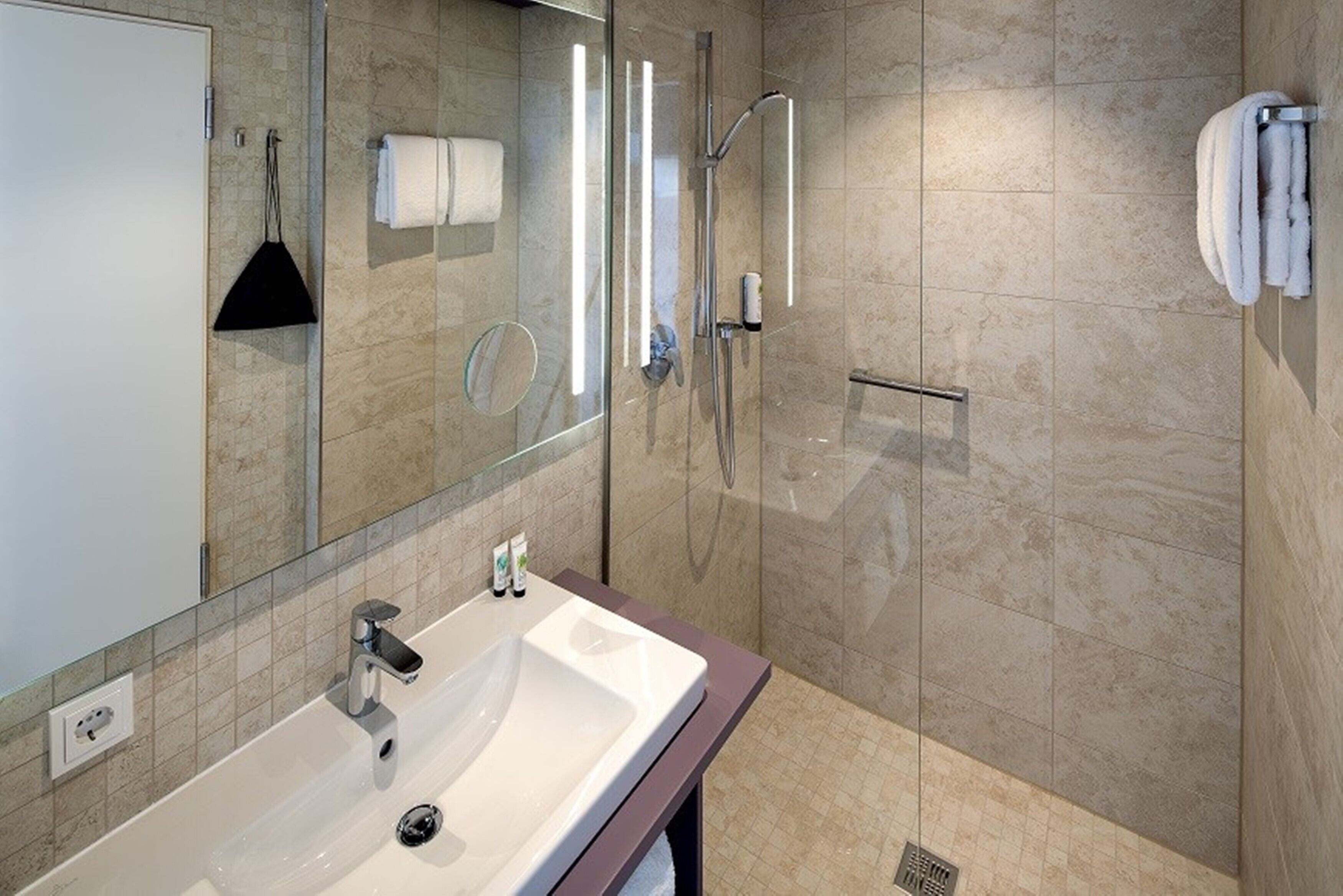A modern bathroom with a large mirror, a white sink, a glass-enclosed shower, and beige tiled walls.