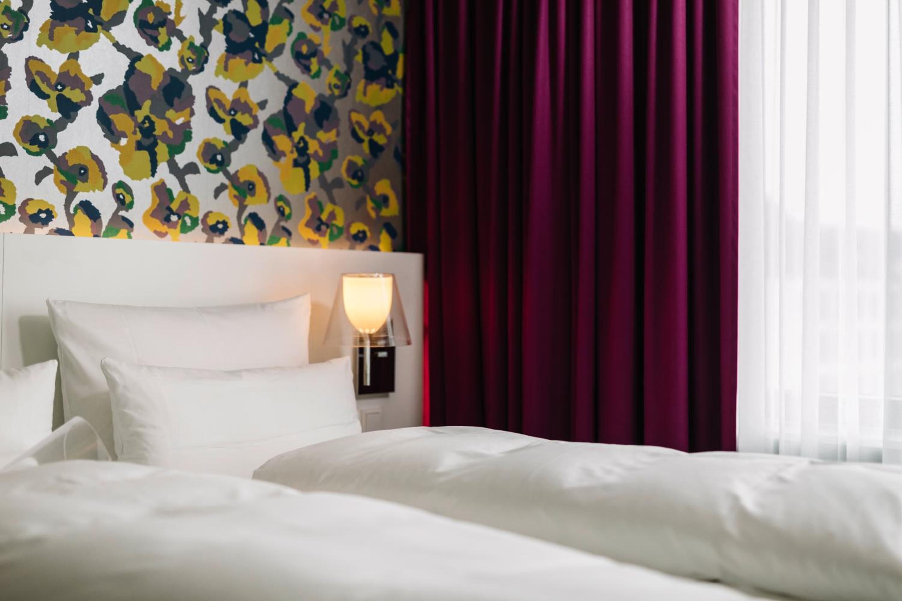 A hotel room with a bed, white bedding, a wall with a floral pattern, a nightstand with a lamp, and purple curtains.